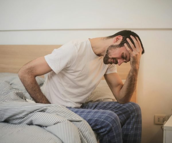 A young man in pajamas holding his head, sitting on a bed, appears to be experiencing a headache.